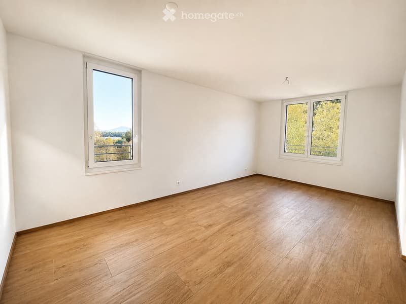 Empty room with wooden flooring and white walls, featuring two windows with white frames and a view of greenery outside.