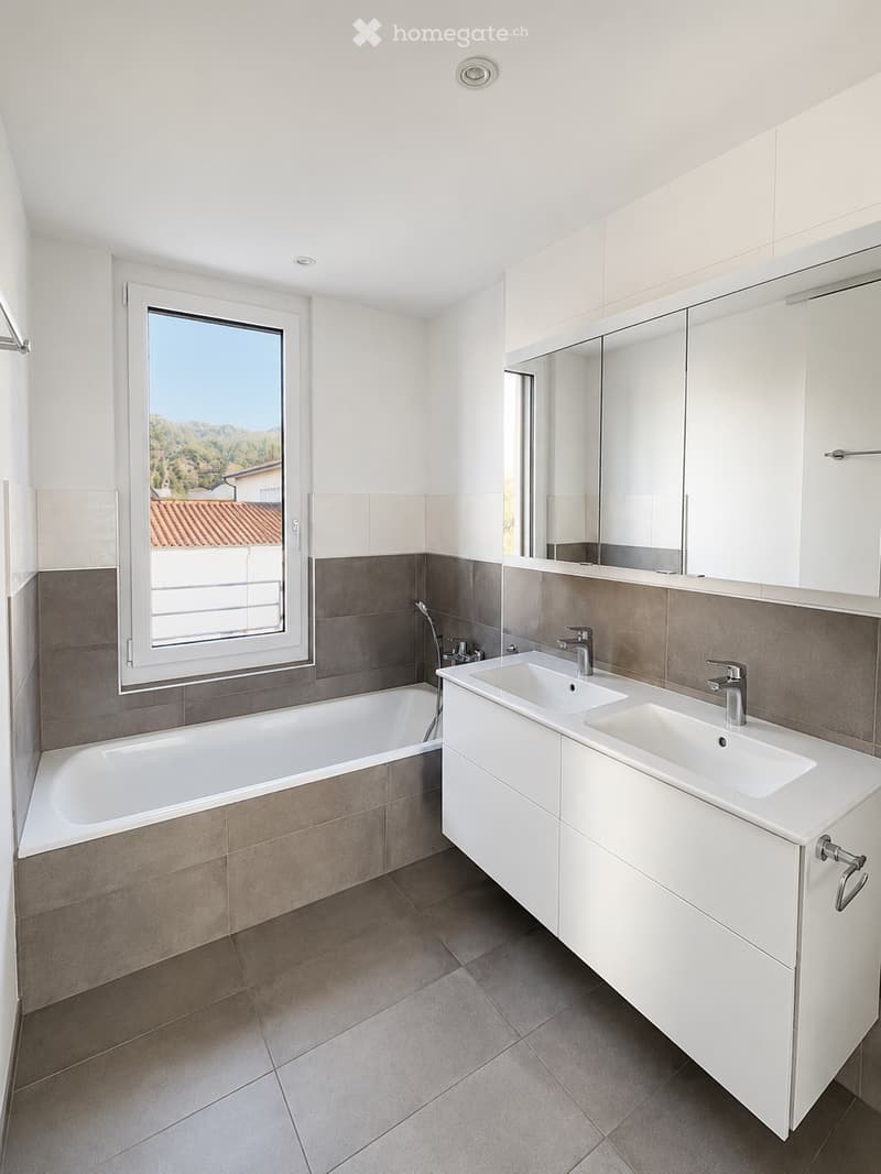Modern bathroom with a bathtub, double sink, large mirror, and a window offering a scenic view.