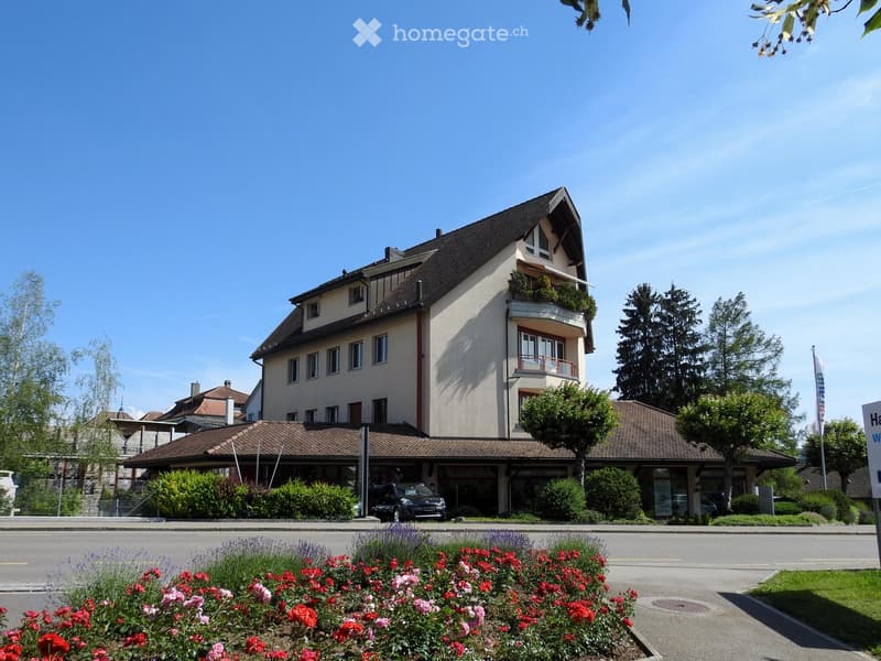 Three-story house, brown roof, beige walls, large windows, car parked in front, plants, trees, street sign, fence, flowers, grassy area