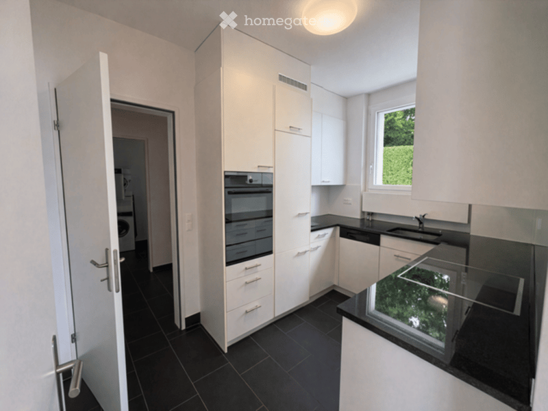 Modern kitchen with white cabinets, black countertops, and a built-in oven. Features a window with a view of greenery and a door leading to another room.