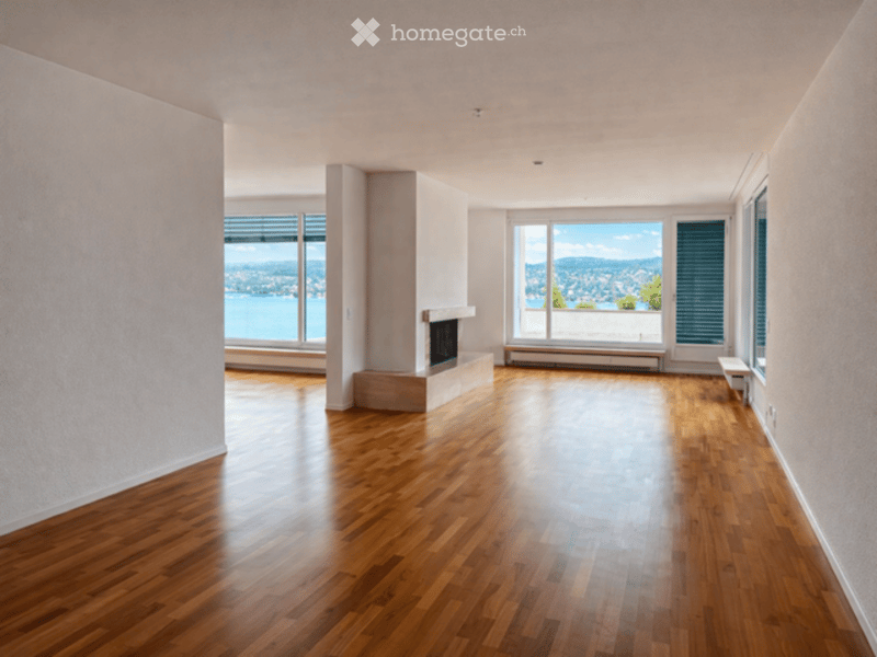 Empty room with polished hardwood floors, large windows offering a scenic view, and a fireplace with a built-in bench.
