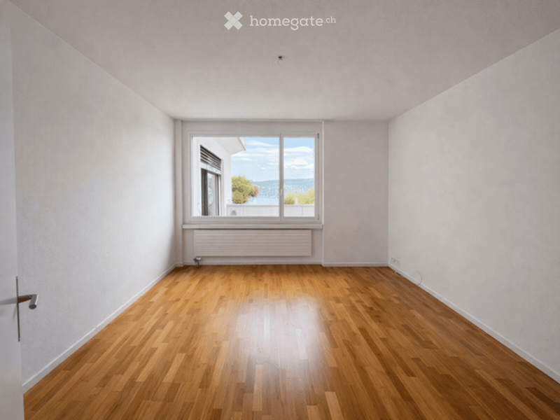 Empty room with wooden flooring, white walls, and a large window offering a scenic view.