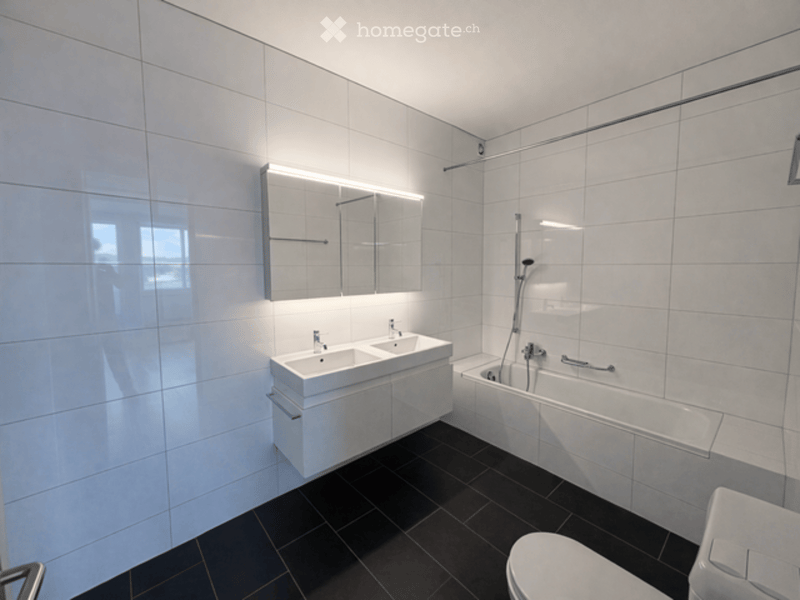 Modern bathroom with white tiles, black floor, double sink, bathtub, toilet, and mirrored cabinet.