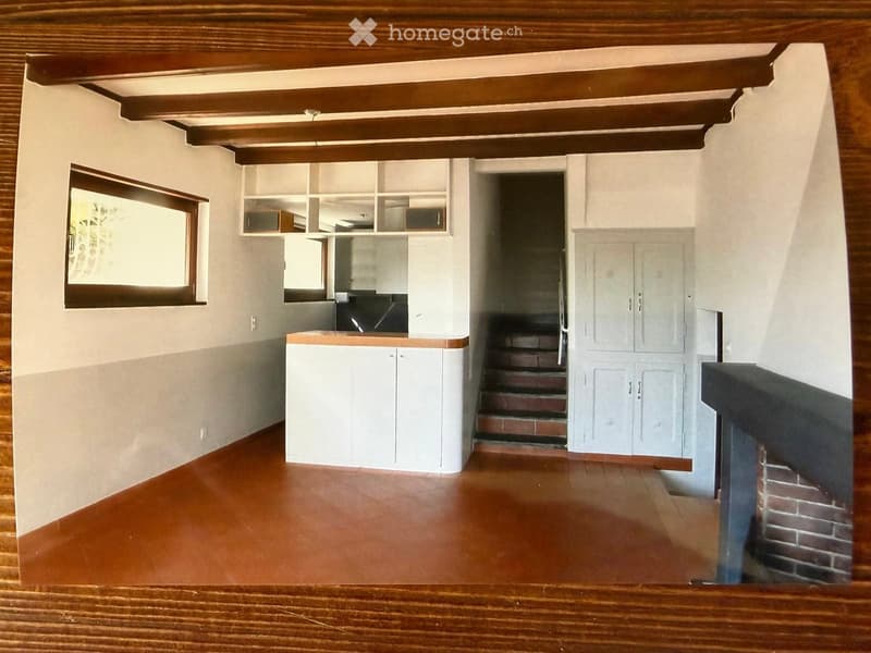 Empty room with wooden ceiling beams, white walls, and a wooden floor. Features include built-in cabinets, a window, and a staircase leading to another level.