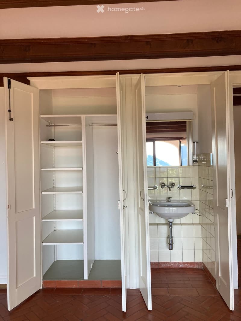 Bathroom with a white sink, mirror, and shelves in an adjacent closet. The bathroom has tiled walls and a red brick floor.