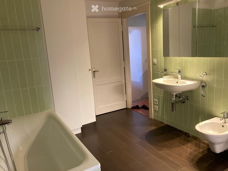 Modern bathroom with green tiled walls, dark wooden floor, bathtub, sink, toilet, and mirror. A door leads to another room.