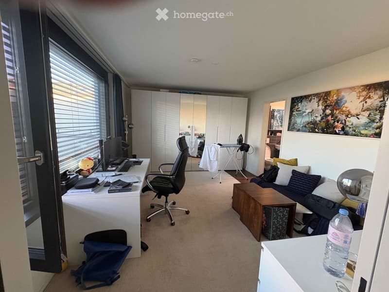 A furnished room with a desk, chair, sofa, and a small ironing board in the corner. The room features large windows with blinds, a water bottle on a small table, and a colorful painting on the wall.