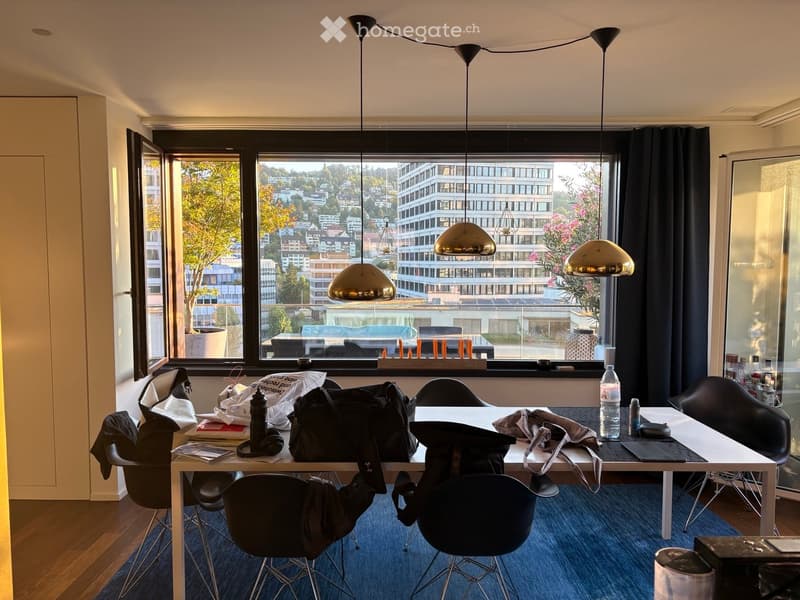 Modern office space with large windows offering a city view, a white table surrounded by black chairs, and various personal items including a backpack, laptop, and water bottle.