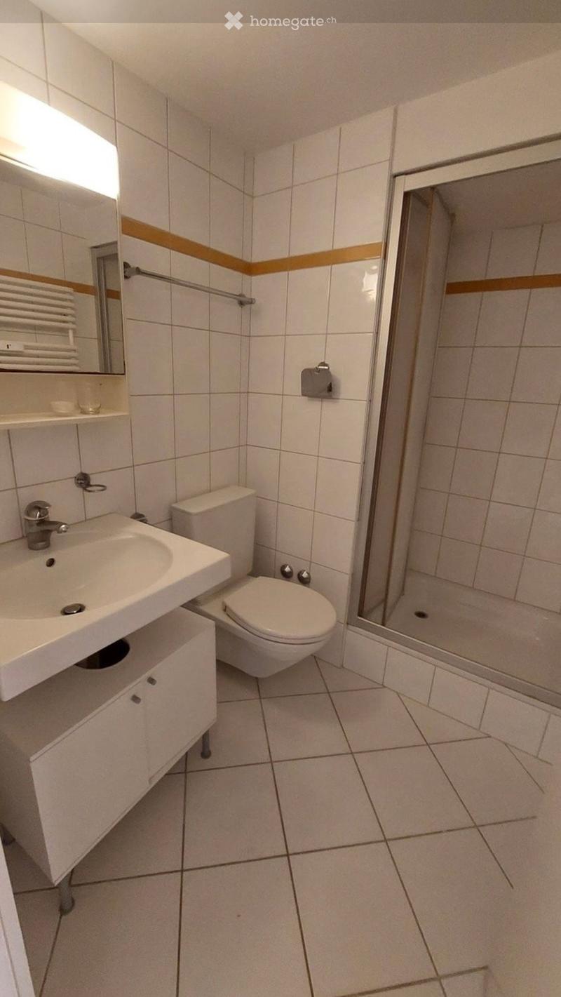 Modern bathroom with white tiles, a sink, toilet, mirror, and a glass-enclosed shower. Features a towel rack and a small shelf with folded towels.