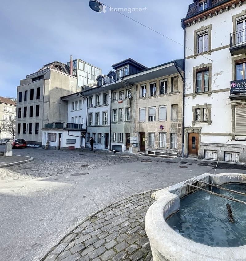 A cobblestone square with a stone fountain, surrounded by multi-story residential buildings with varied architectural styles and windows.