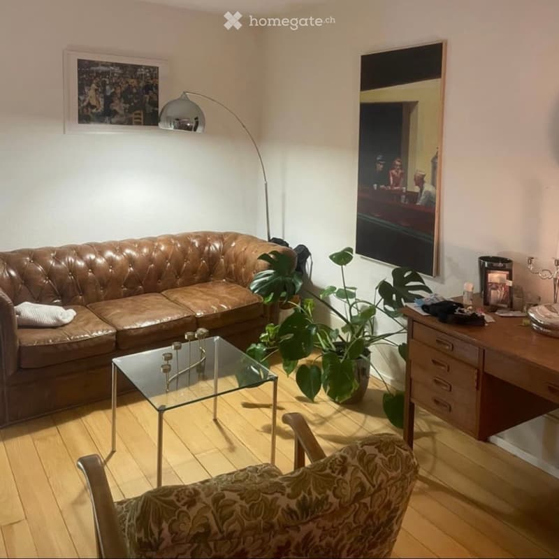 Living room with a brown leather sofa, glass coffee table, patterned armchair, floor lamp, potted plant, and a wooden cabinet with various items on top.