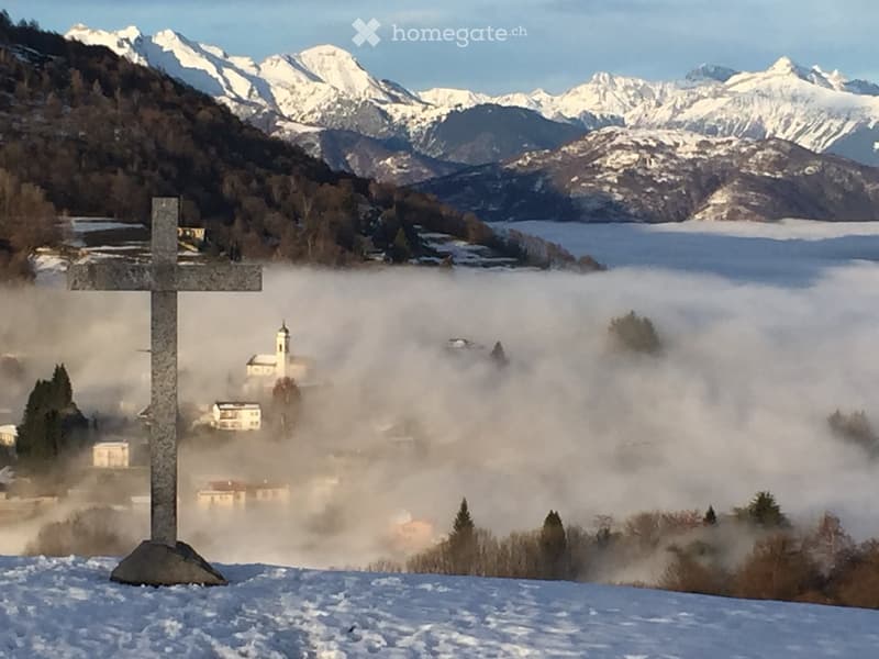 A serene outdoor scene featuring a stone cross in the foreground, with a small village partially obscured by mist in the background, surrounded by snow-capped mountains and a calm lake.