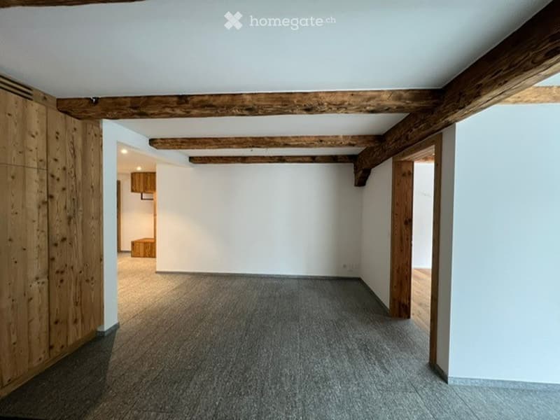 Empty room with exposed wooden beams on the ceiling, grey carpeted floor, white walls, and wooden paneling on one side. There are doorways leading to other rooms.