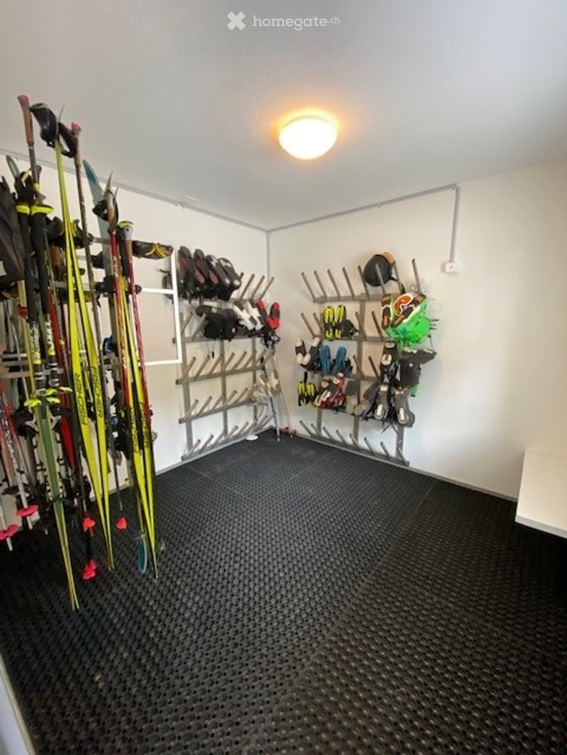 A small, organized storage room with a black rubber floor and white walls. It features a ski rack holding multiple pairs of skis, ski poles, a snowboard, boots, helmets, and gloves neatly arranged on hooks and shelves.