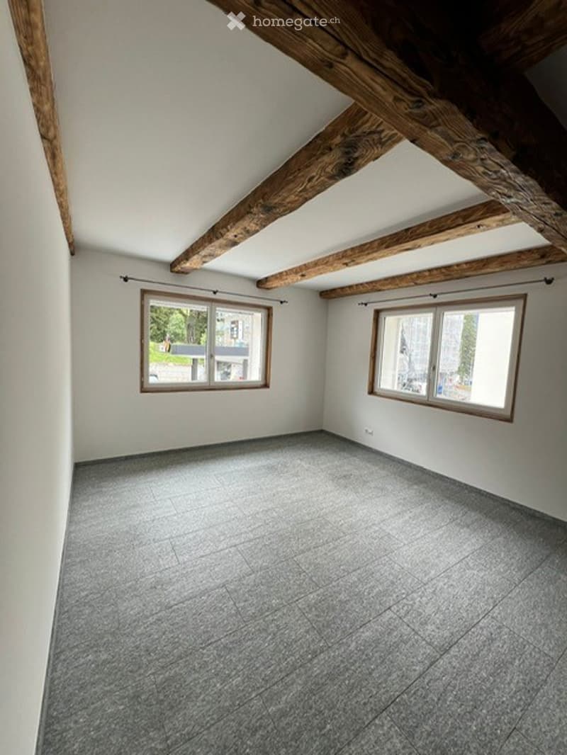Empty room with exposed wooden beams, grey tiled floor, and two windows with wooden frames.