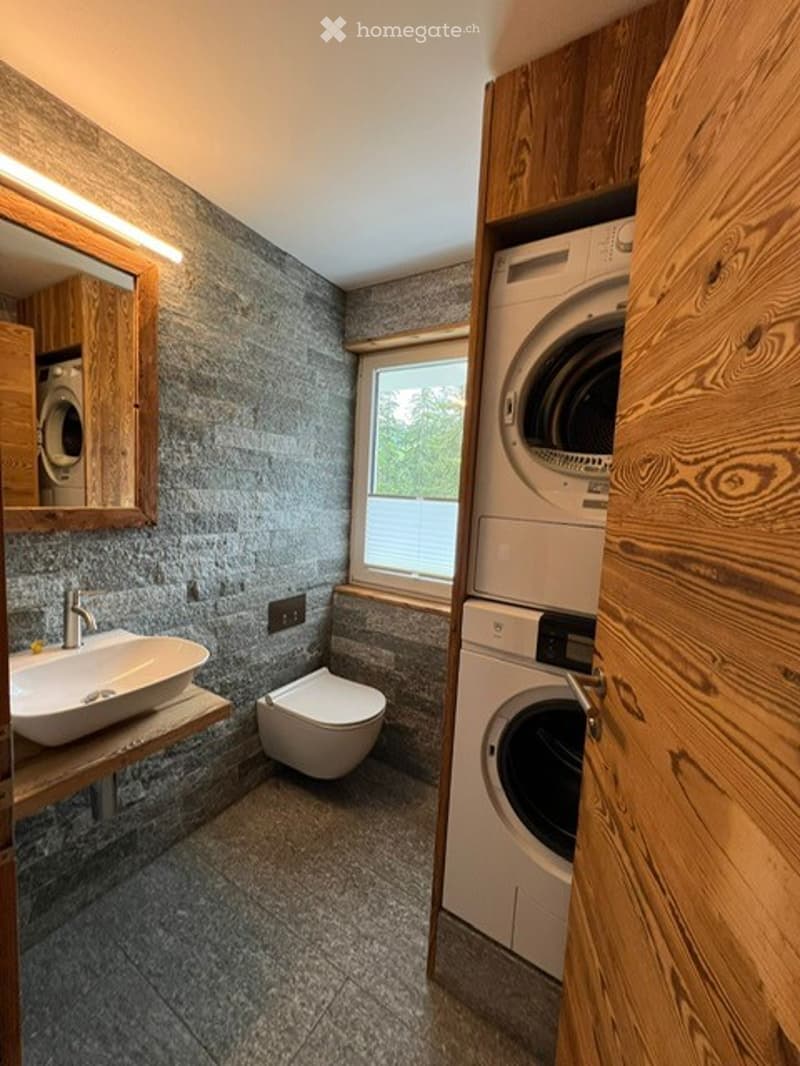 Modern bathroom with grey stone walls, a sink, toilet, mirror, and a washing machine in a wooden cabinet.