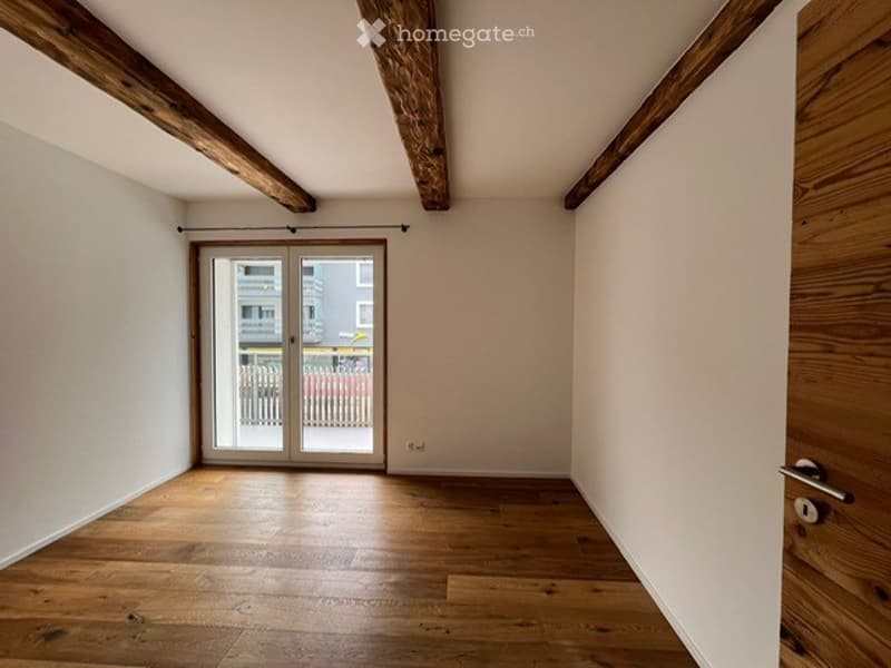 Empty room with wooden floor, exposed wooden beams, large glass sliding door leading to a balcony, and white walls.
