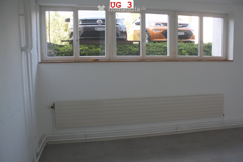 Empty room with white walls, large windows showing parked cars and greenery, radiator, and a grey floor.