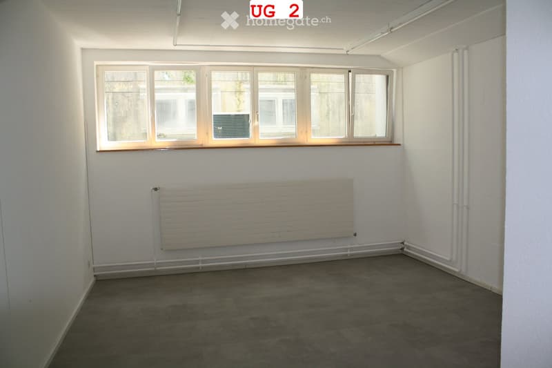 Empty room with white walls, grey tiled floor, large window with wooden frame, radiator, and visible pipes along the wall.