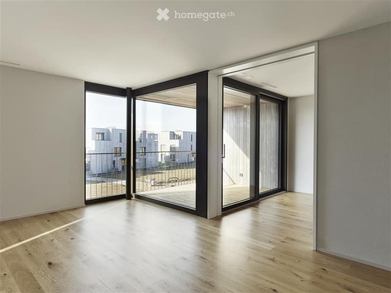 Empty room with light wooden flooring, large sliding glass doors leading to a balcony, and white walls.