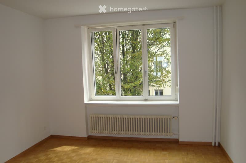 Empty room with wooden flooring, white walls, large window with white frame, and a radiator below the window.