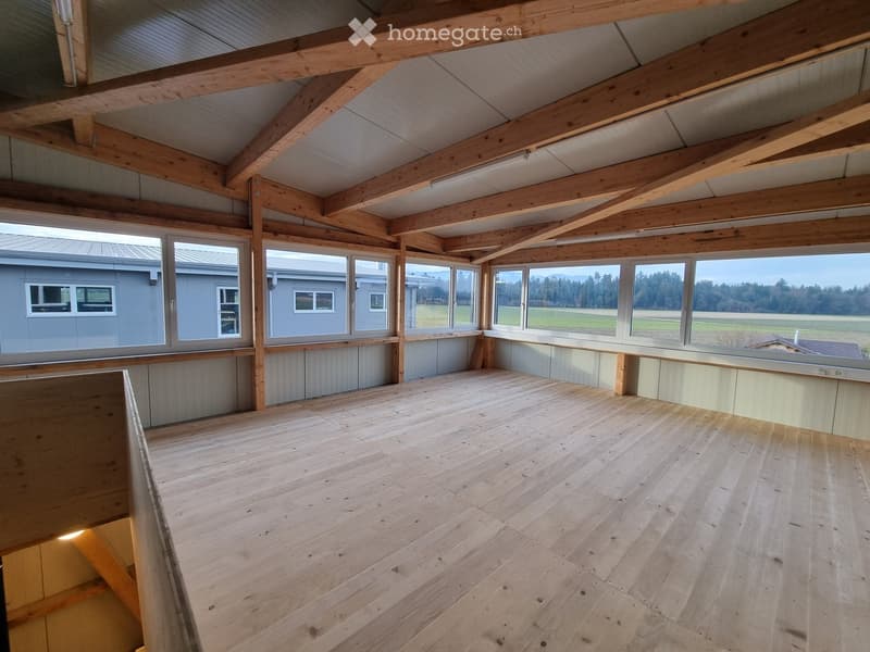 Empty room with wooden floor and exposed wooden beams, large windows offering a view of the landscape.