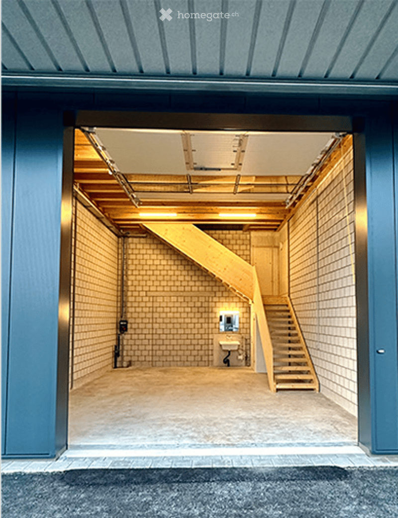 Modern garage with a wooden staircase leading to an upper level, tiled walls, and a small bathroom area visible.