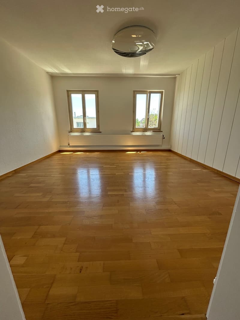 Empty room with wooden flooring, white walls, and two windows with white frames. A ceiling light fixture is visible.