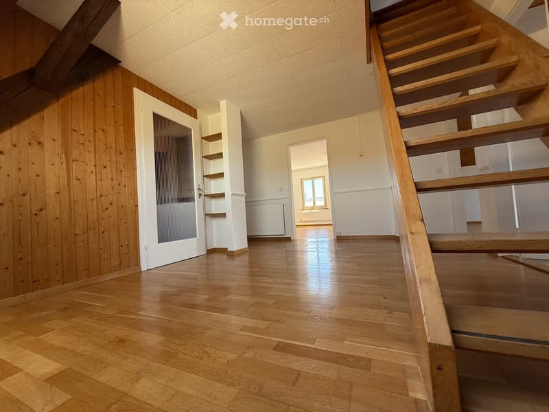Bright hallway with wooden flooring, white walls, and a staircase leading upwards. Features a glass-paneled door and built-in shelving unit.