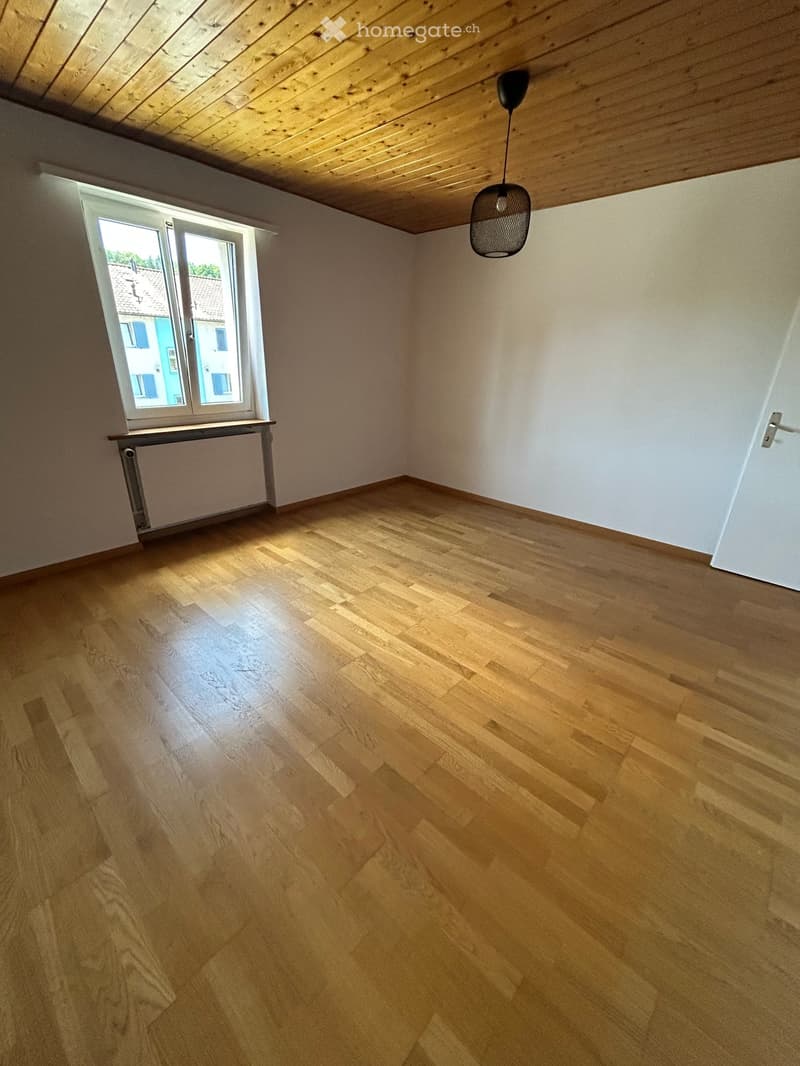 Empty room with wooden flooring, white walls, a window with a radiator below, and a hanging light fixture.