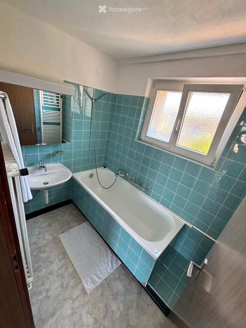 Bathroom with blue tiled walls, a bathtub, a sink, a mirror, and a radiator. The room has a window and a towel hanging on the side.