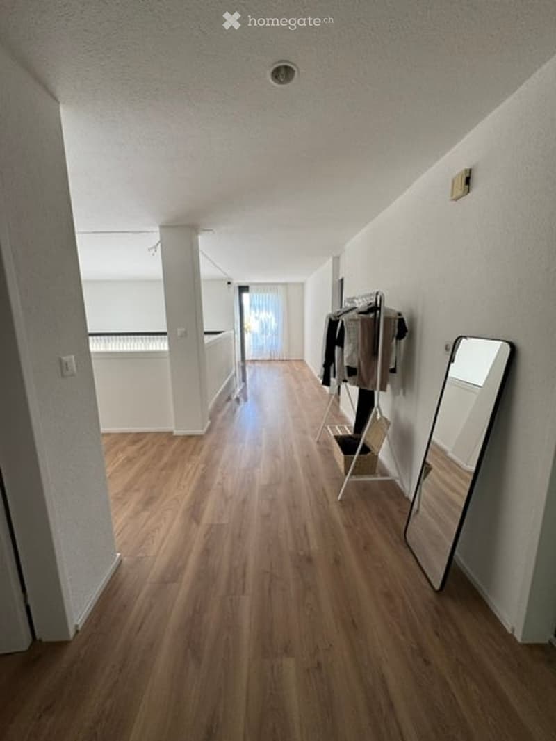 Bright hallway with wooden flooring, white walls, and a coat rack with jackets. A large mirror is placed on the right wall, and a balcony is visible at the end of the hallway.