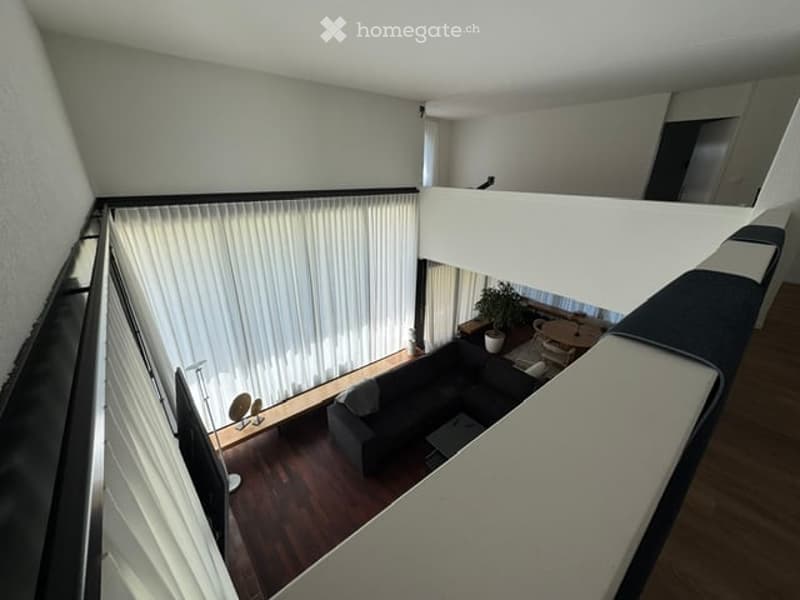Modern, open-concept living space with a dark grey sofa, a small round table with chairs, a potted plant, and a floor lamp near the window with white blinds. The room features wooden flooring and a raised platform with a railing.