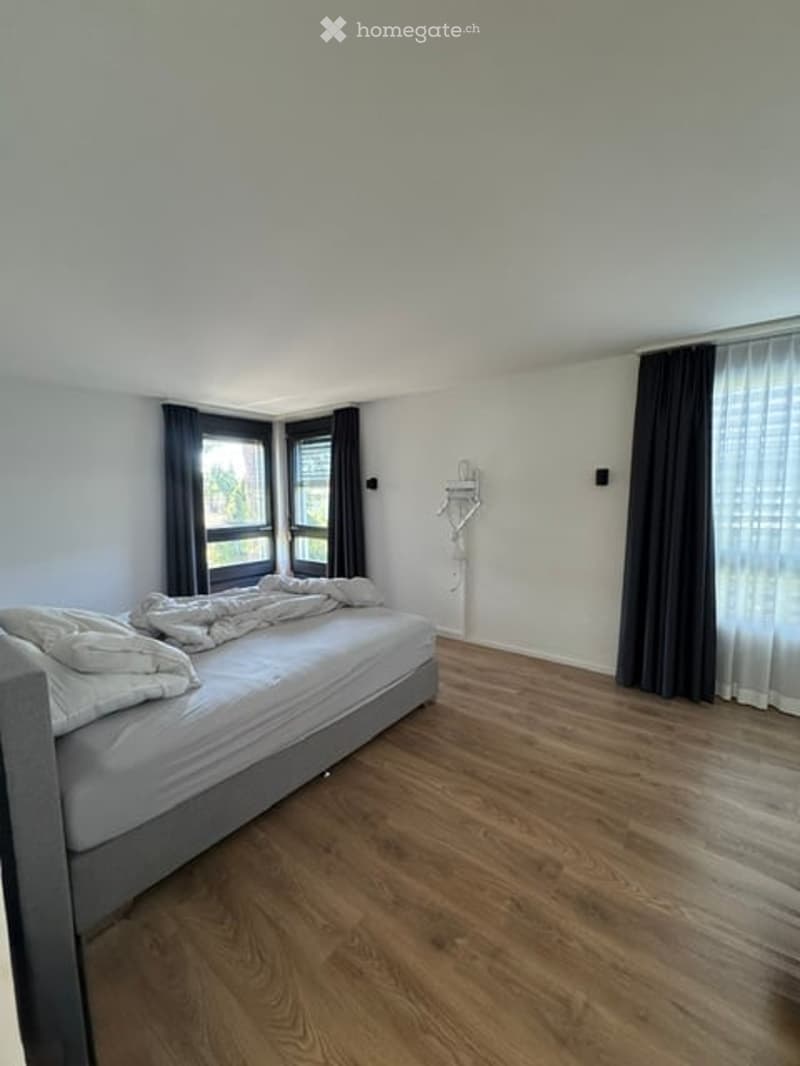 Minimalist bedroom with a single bed, light grey bedding, wooden flooring, and dark curtains on the windows.