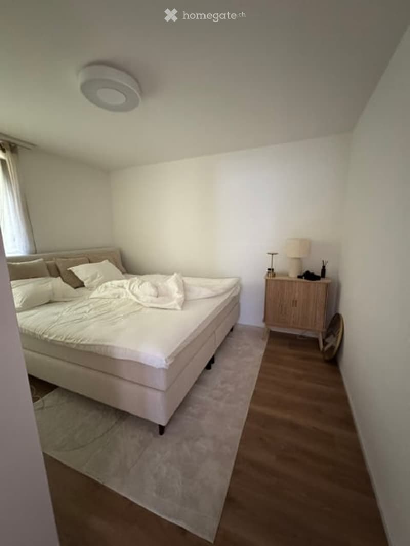 Modern bedroom with a large bed, white bedding, a nightstand with a lamp, and a mirror on the wall. The room features light-colored walls, wooden flooring, and a large window with curtains.
