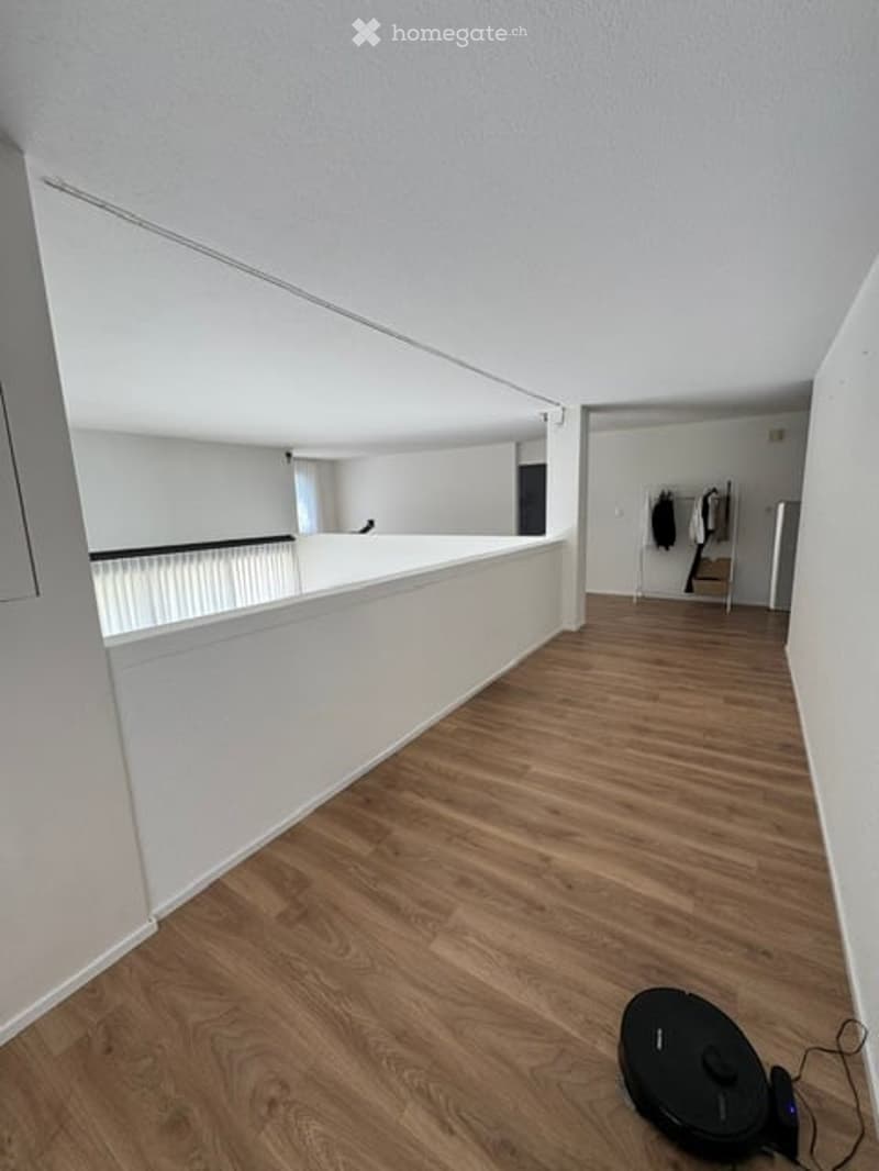 Empty room with wooden flooring and white walls, featuring a white railing and a small black robotic vacuum cleaner on the floor.