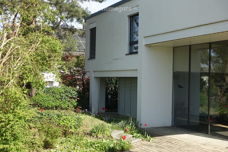 Modern white house with large glass sliding doors, raised ground floor, and well-maintained garden with flowers and shrubs.