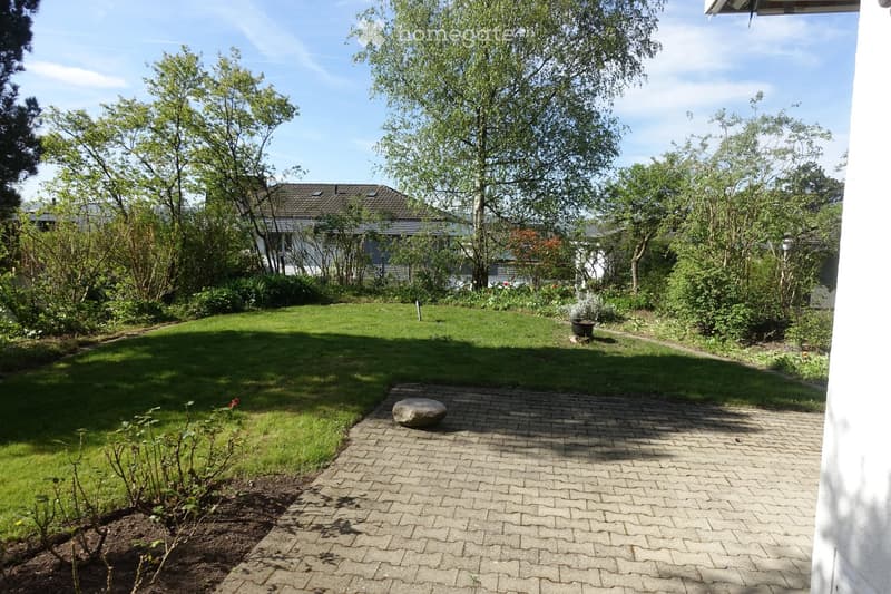 Well-maintained garden with a paved pathway, green lawn, trees, and a house in the background.