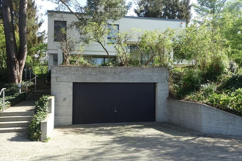 Modern two-story house with a flat roof, large windows, and a concrete garage entrance. The house is surrounded by lush greenery and trees, with a raised ground floor and steps leading up to the entrance.