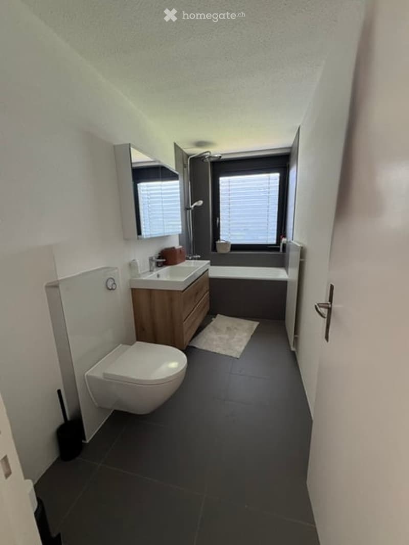 Modern bathroom with dark tiled floor, white toilet, wooden sink cabinet, bathtub, and large window with blinds.