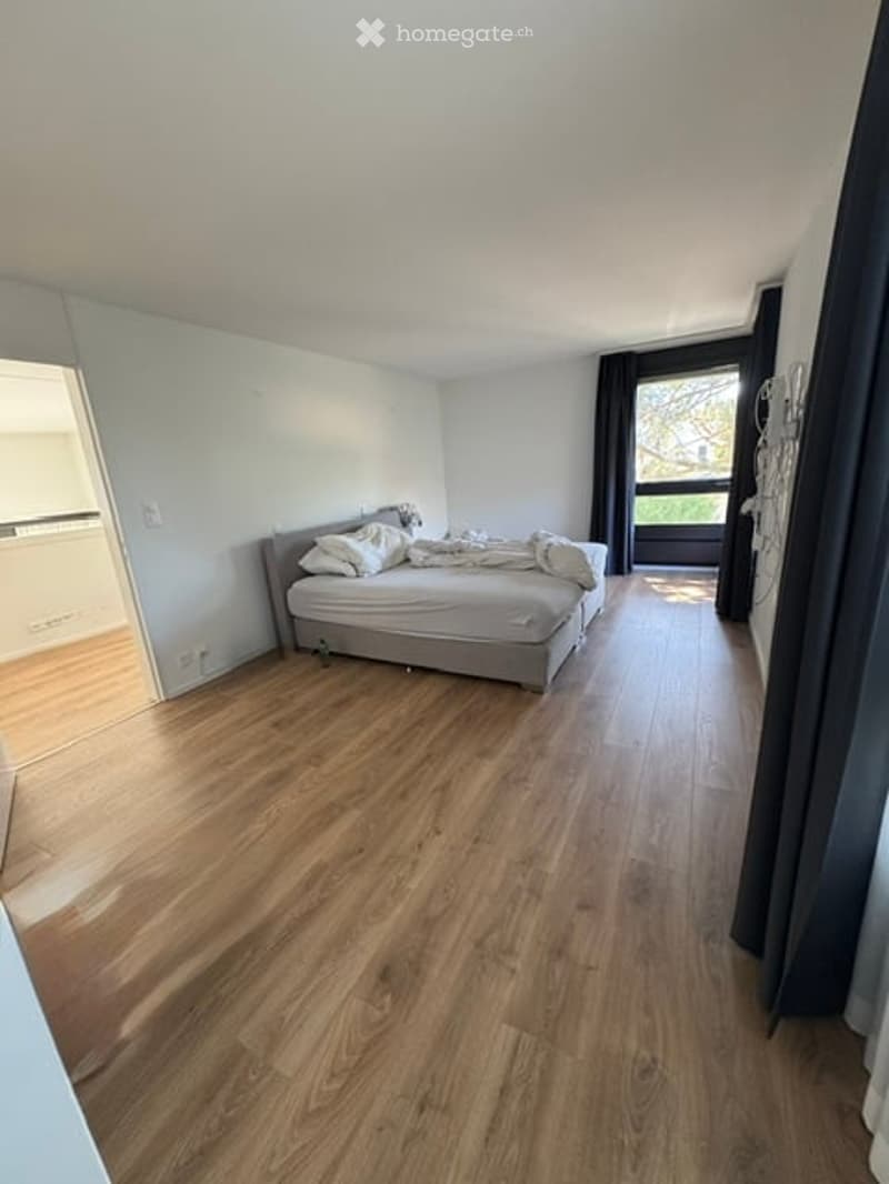 Bright bedroom with wooden flooring, a grey bed with white pillows and a blanket, and large windows with dark curtains.