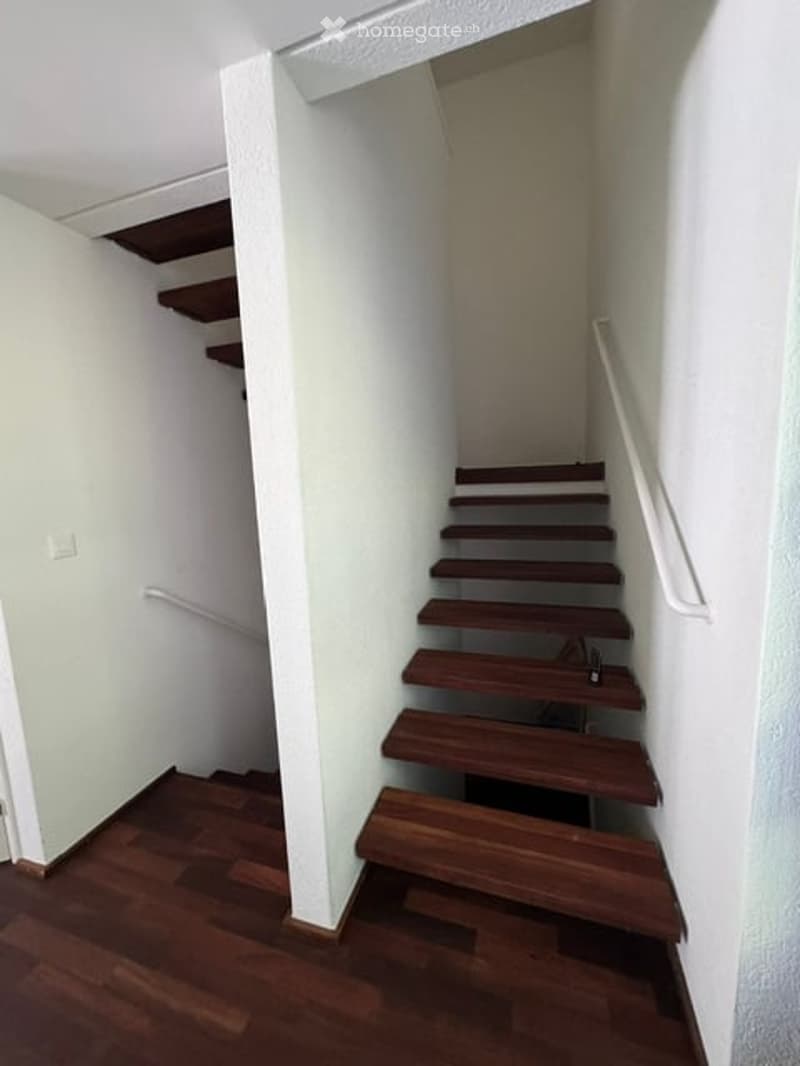 A narrow staircase with wooden steps and a white handrail, leading up to a landing area. The walls are painted white, and the floor is made of polished wood.