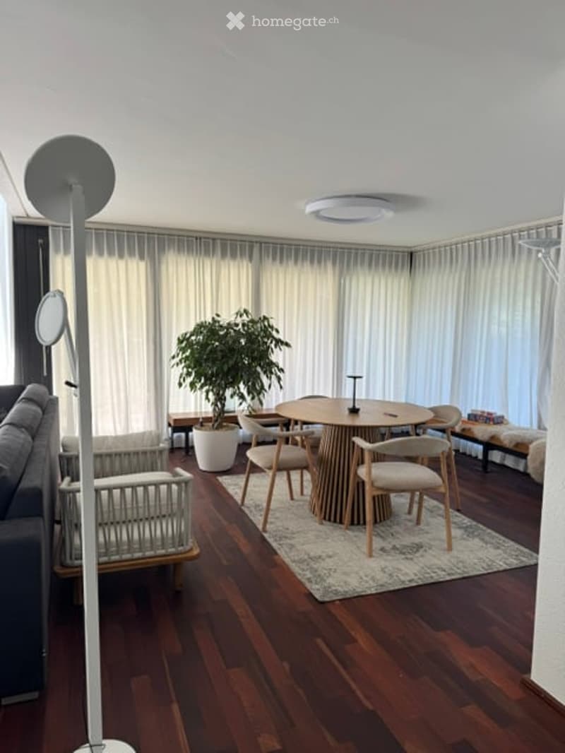 Modern living room with wooden flooring, large windows with white curtains, a round dining table with chairs, a grey sofa, and a floor lamp.