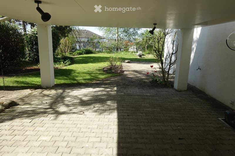 A covered outdoor area with a brick pathway leading to a well-maintained lawn and garden, featuring trees, shrubs, and a small water fountain.