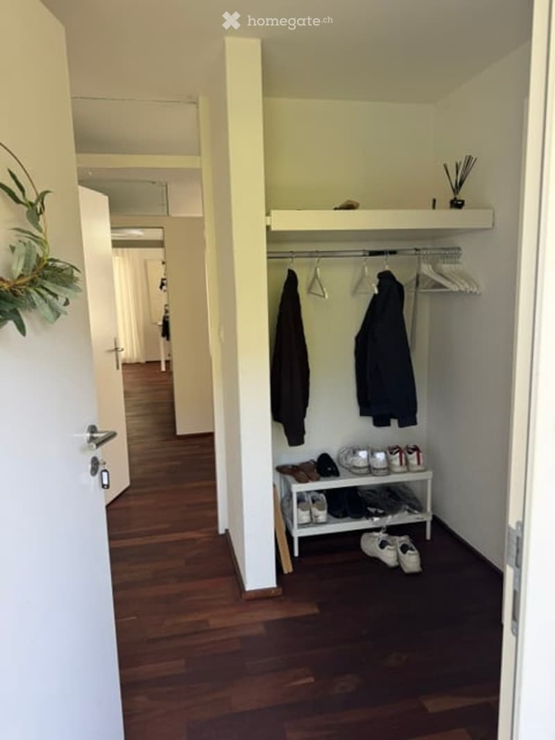 A narrow hallway with wooden flooring, white walls, and a coat rack with hanging coats and shoes. A small shelf holds additional shoes and accessories.
