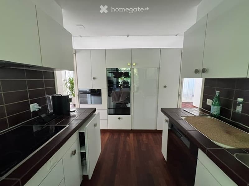 Modern kitchen with white cabinets, dark countertops, and wooden flooring. Features include a sink, oven, coffee maker, and a bottle on the counter.