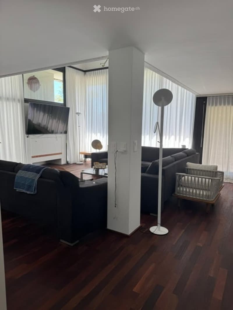Modern living room with dark wooden flooring, large windows with white curtains, two sofas, coffee table, floor lamp, television, and ottoman.