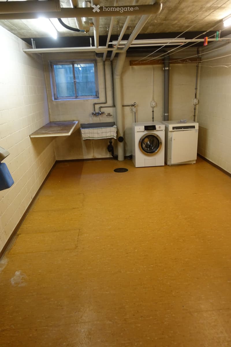 Laundry room with a washing machine, sink, and small table, concrete floor, exposed pipes, and a window.