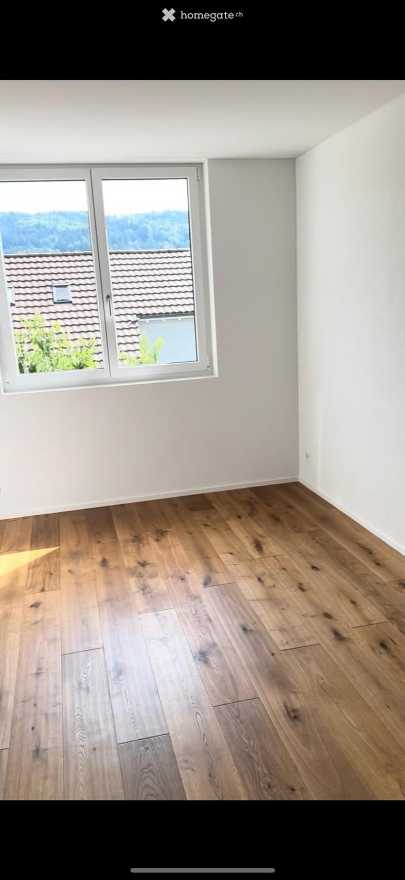 Empty room with wooden flooring and large windows offering a view of the roof and surrounding landscape.