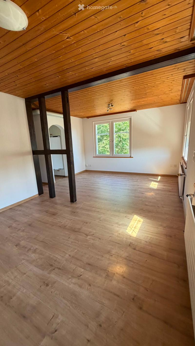 Empty room with wooden floor, exposed wooden ceiling beams, and large windows letting in natural light.